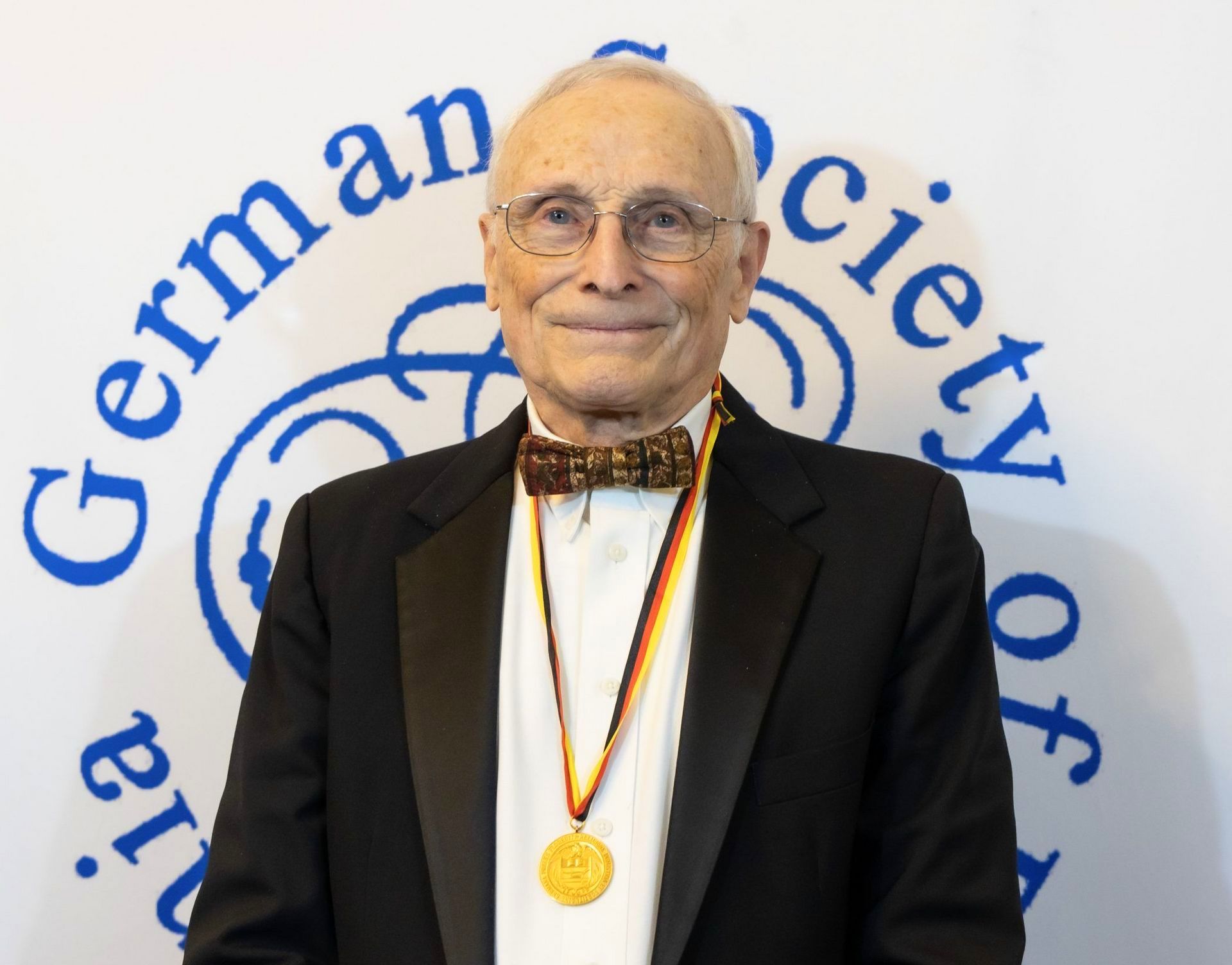 A man in a tuxedo with a medal around his neck is standing in front of a german society logo