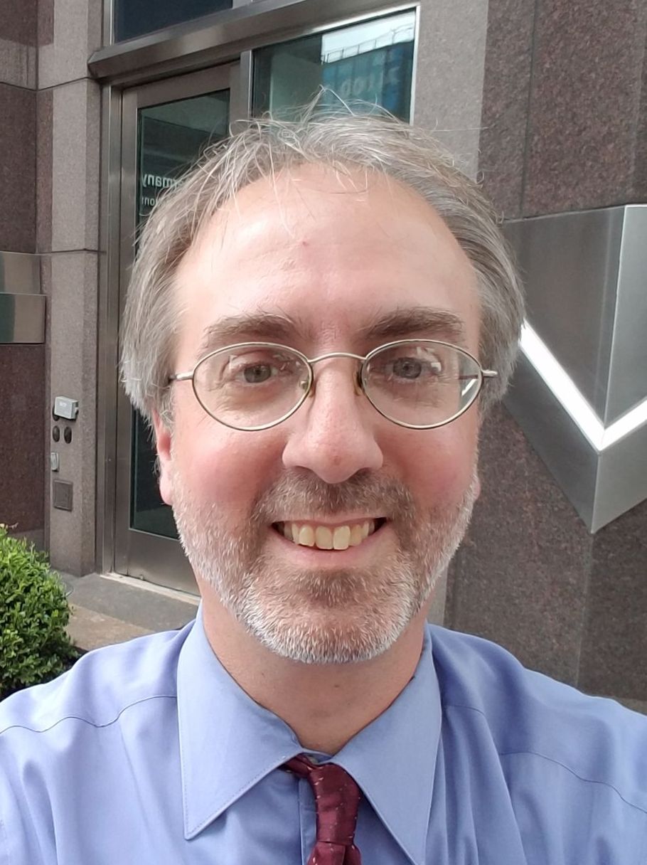 A man with glasses and a beard is smiling in front of a building.