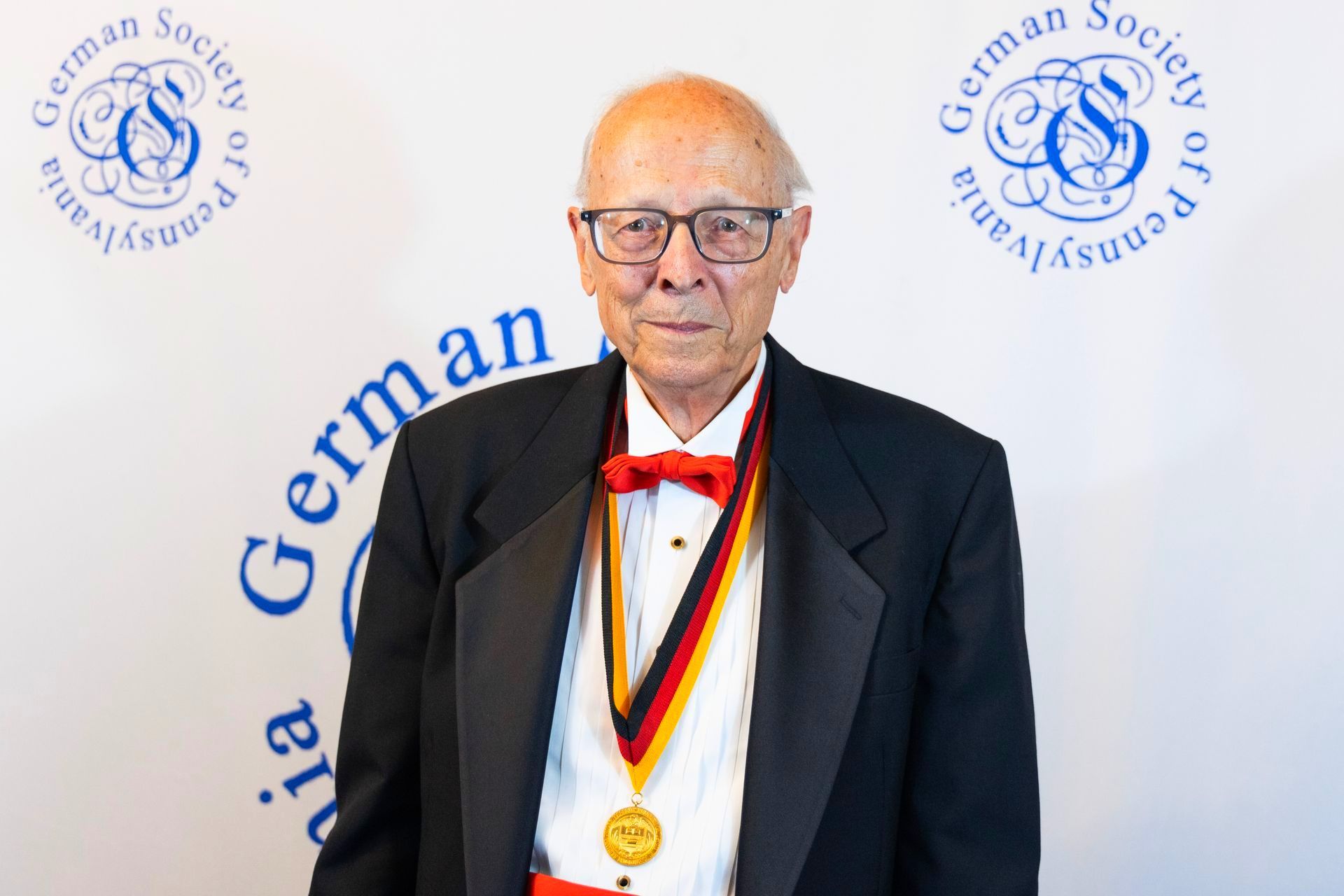 A man in a tuxedo with a medal around his neck is standing in front of a german society logo.