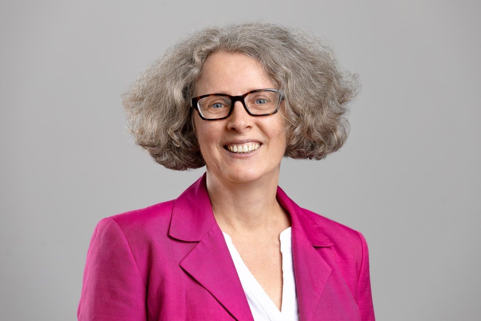 A woman wearing glasses and a pink jacket is smiling.