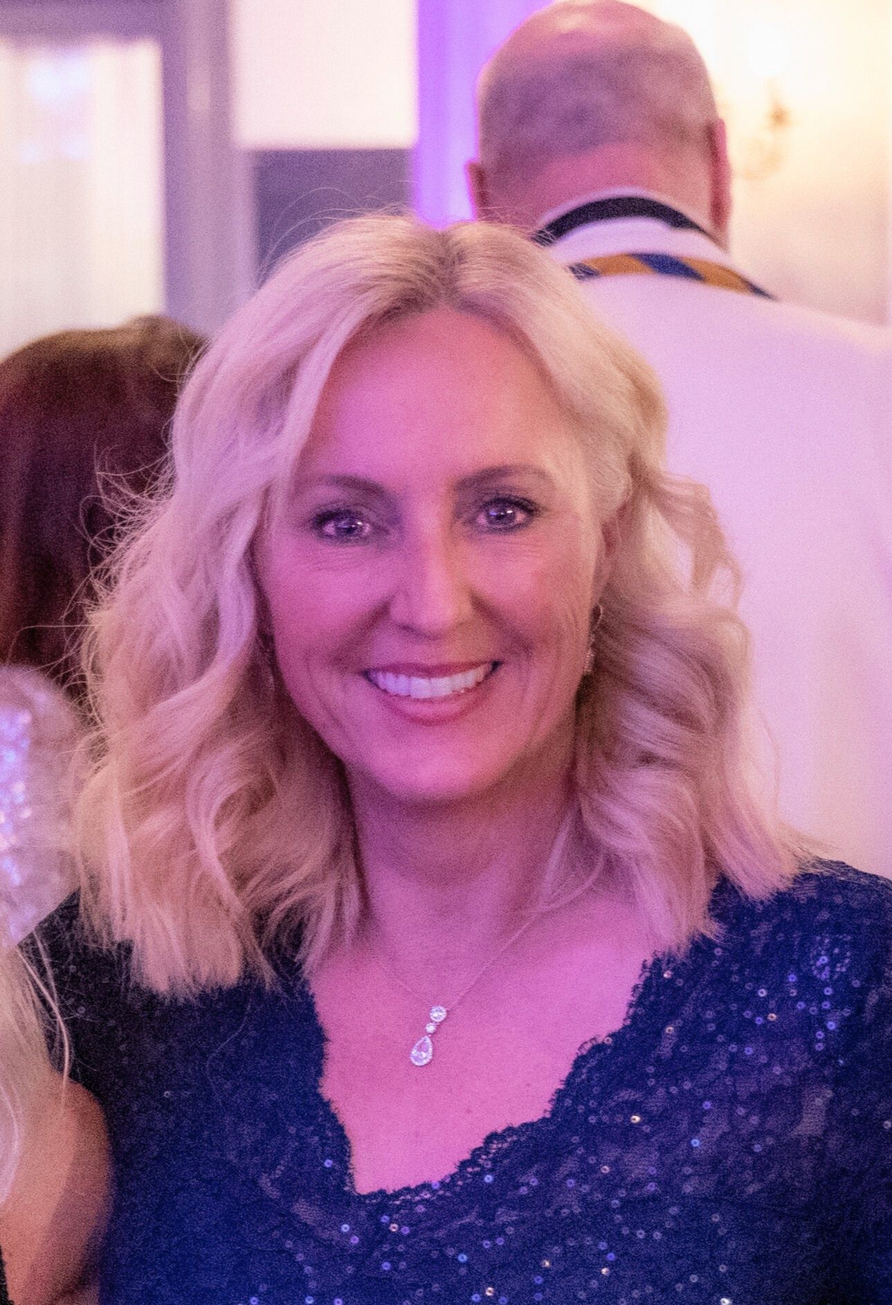 A woman with blonde hair is wearing a blue shirt and a necklace.