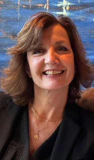 A woman in a black jacket and necklace is smiling in front of a painting.