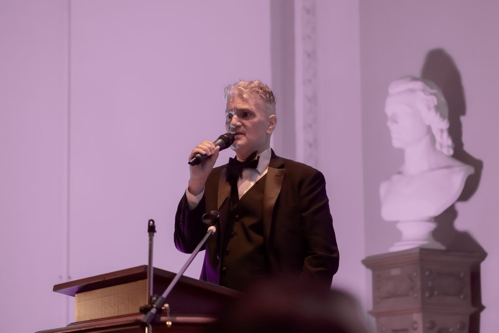 A man in a tuxedo is standing at a podium holding a microphone.