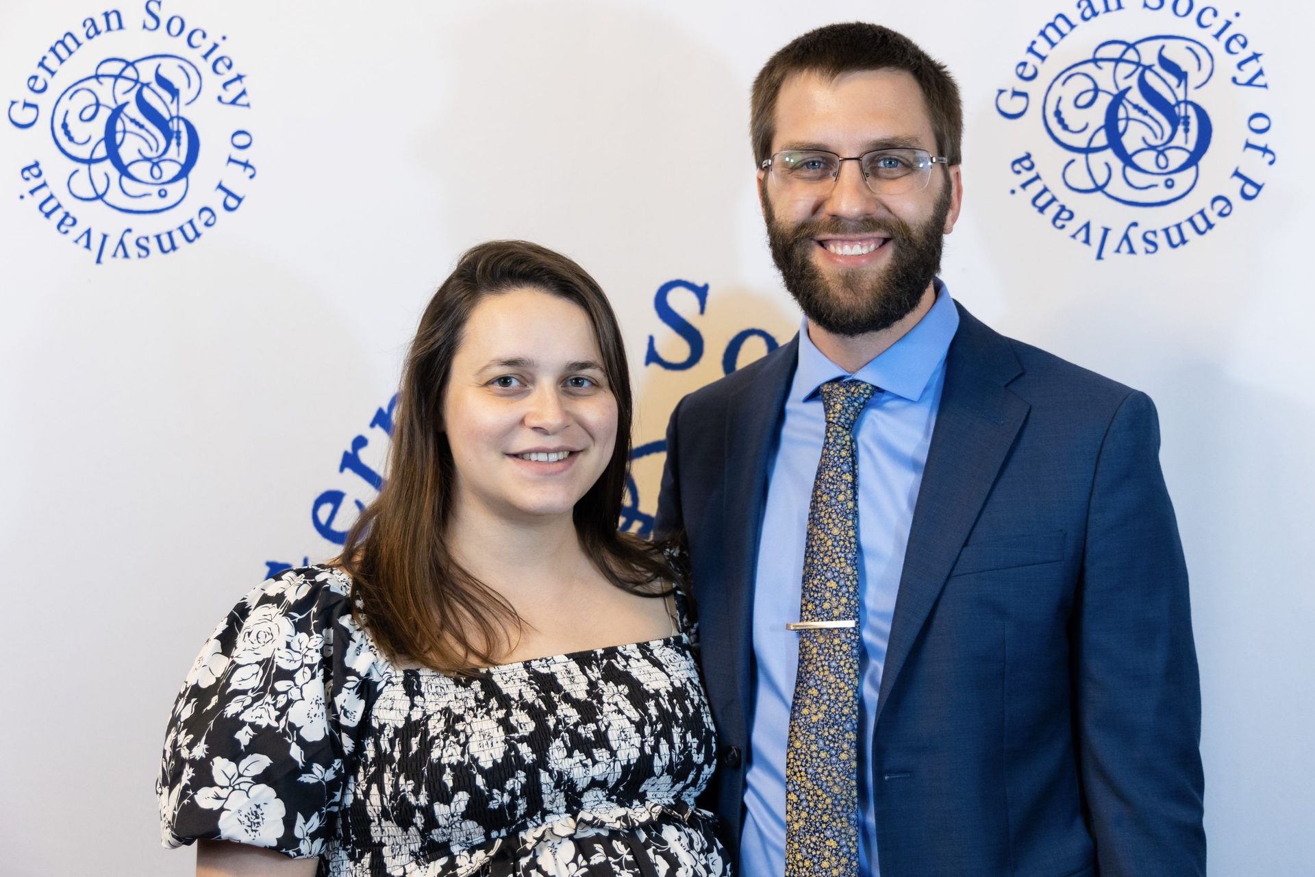 A man and a woman are posing for a picture in front of a german society of pennsylvania logo.