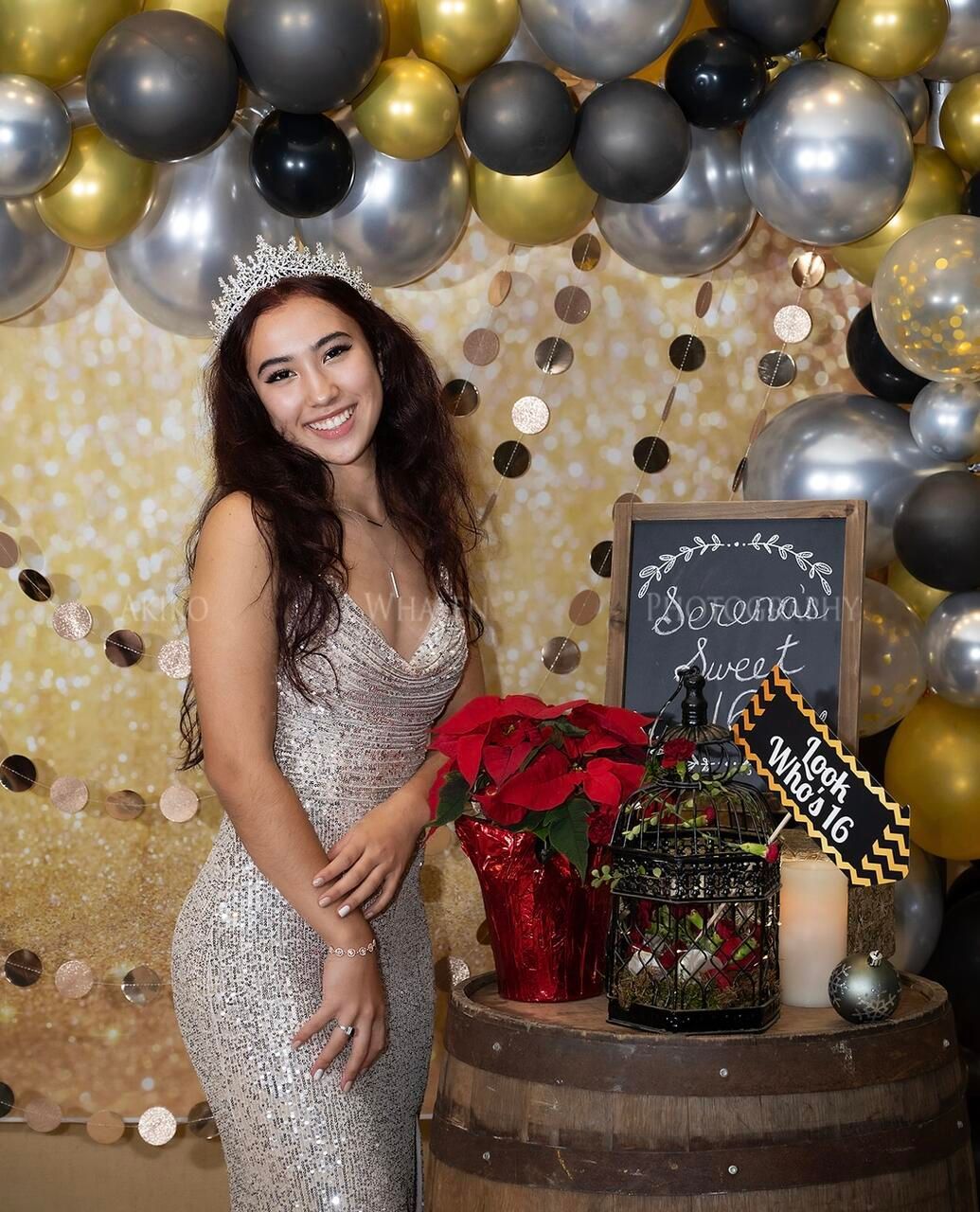 Woman in a sequined dress and tiara smiles at a Sweet 16 party, posing next to a barrel with decor and balloons.
