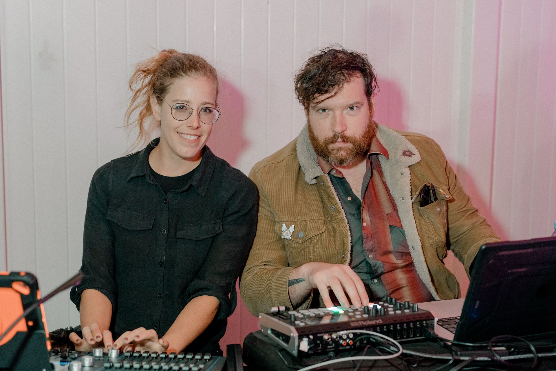 Two people smiling, operating audio equipment at a desk. Woman with glasses, man with beard, in a brightly lit space.