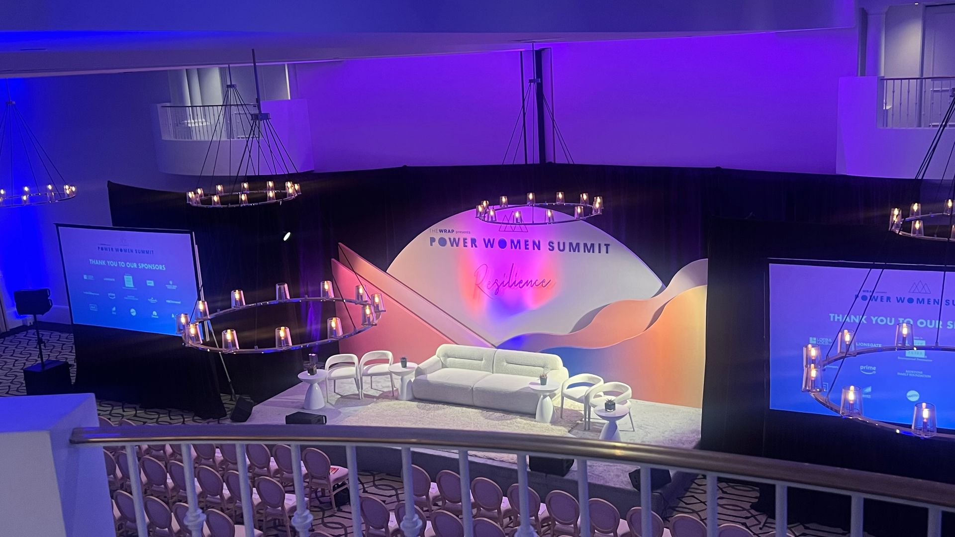 Event stage set up for a summit, featuring a couch, chairs, screens, and decorative lighting. Purple and blue hues.