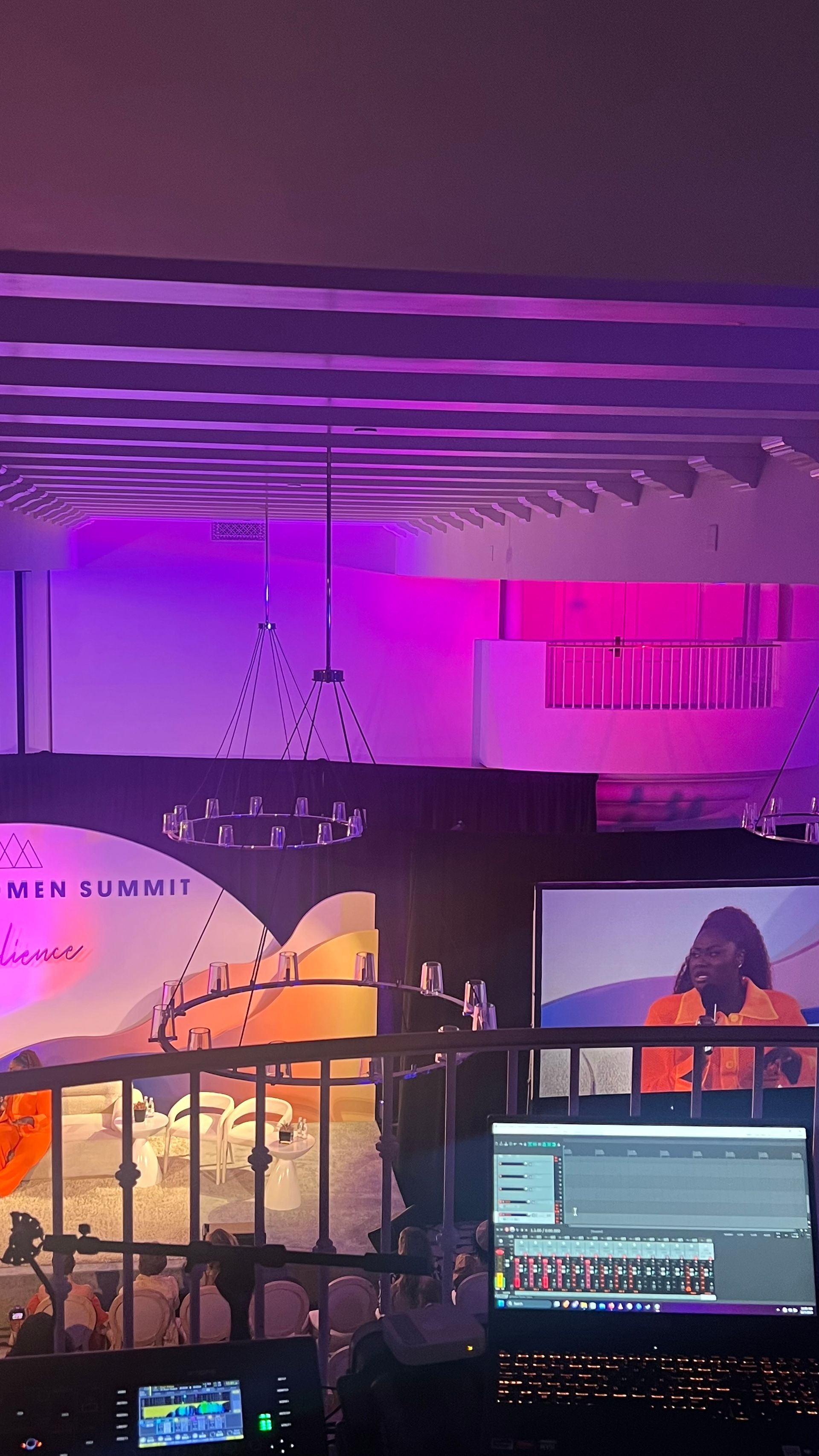 A stage with a speaker and a screen displaying them. Pink and purple lighting illuminates the venue.