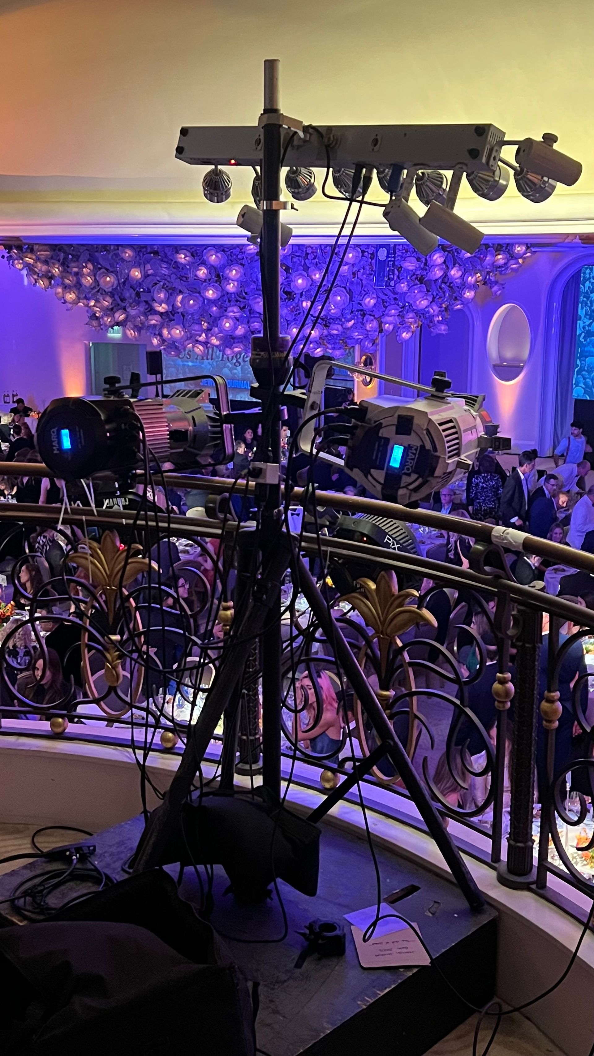 Stage lighting rig set up on a balcony, overlooking a decorated ballroom with people.