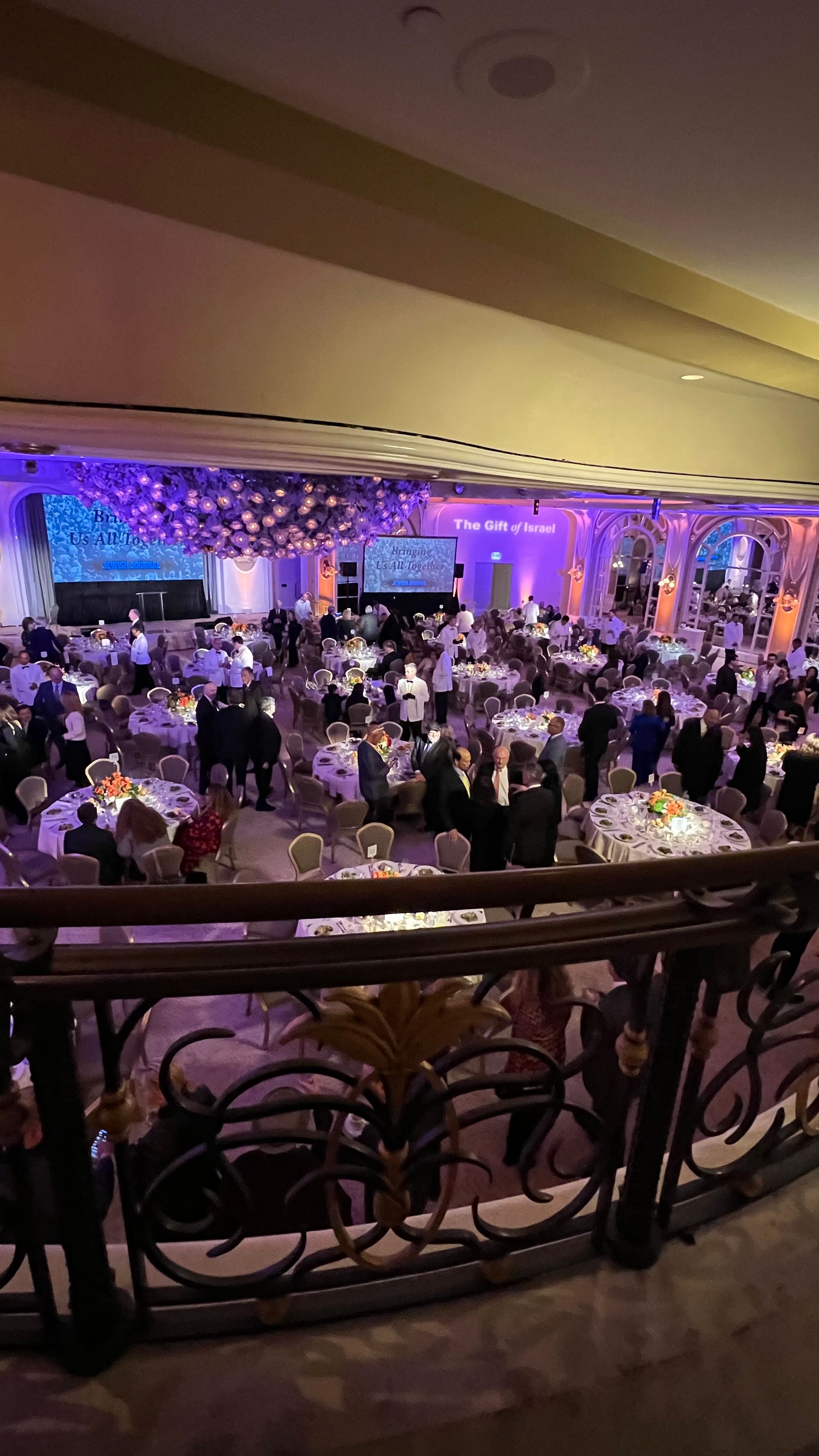 Ballroom with people at tables; purple and orange lighting, decorative balcony in foreground.