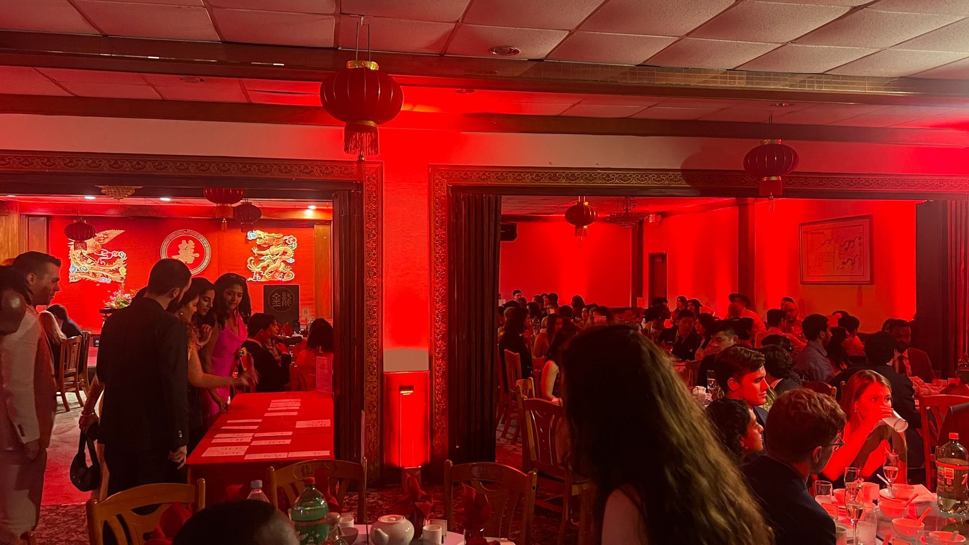 Interior of a banquet hall, lit in red. Many people seated at tables, some socializing. Chinese lanterns hang.
