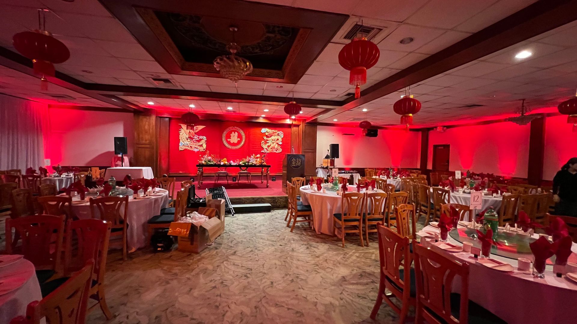 Red-lit Chinese restaurant with tables set for a banquet, stage with floral arrangements, and red lanterns.