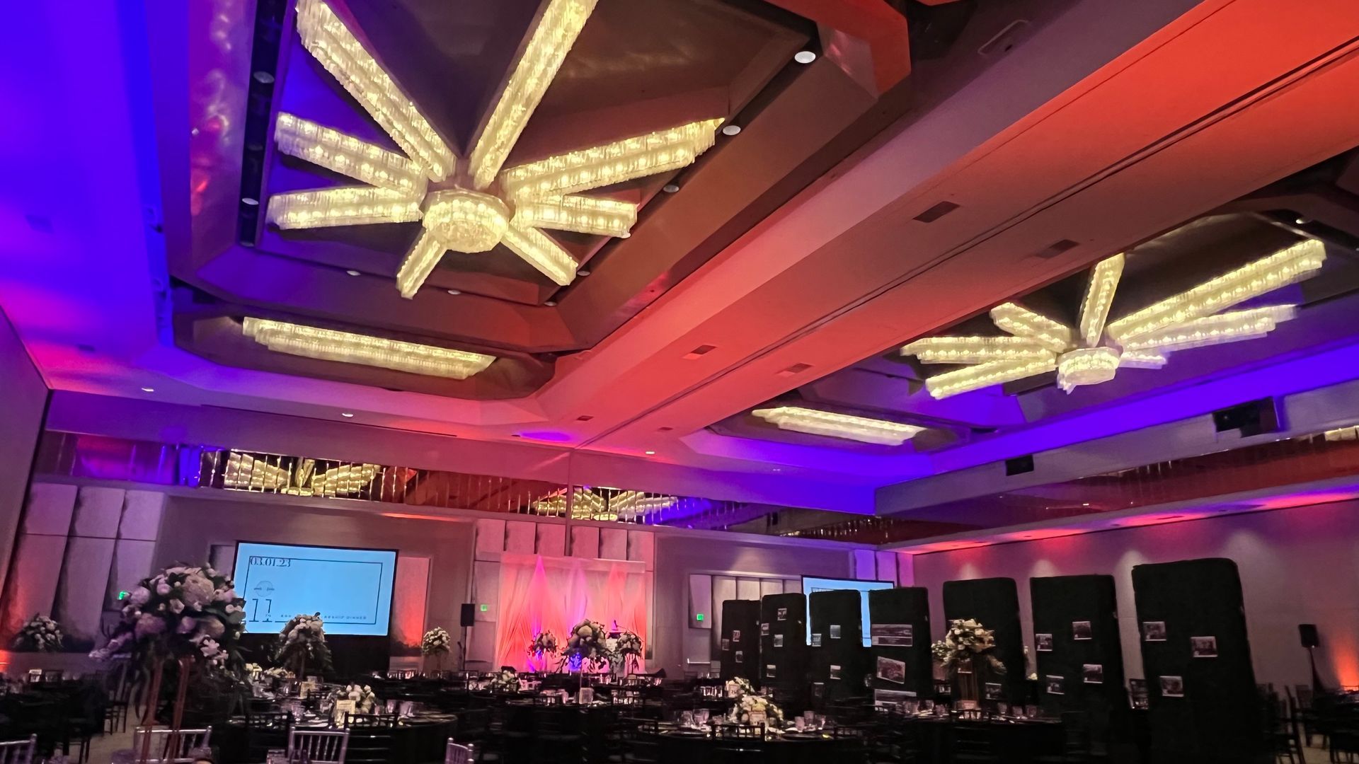 A grand ballroom with decorated ceiling lights, tables set for a banquet, and colorfully lit walls.