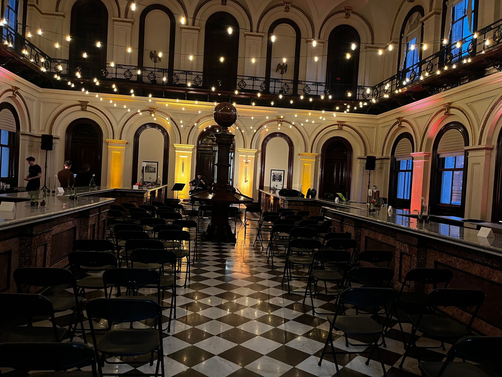 Ornate hall with checkered floor, rows of chairs, and a central sculpture, dimly lit with string lights.