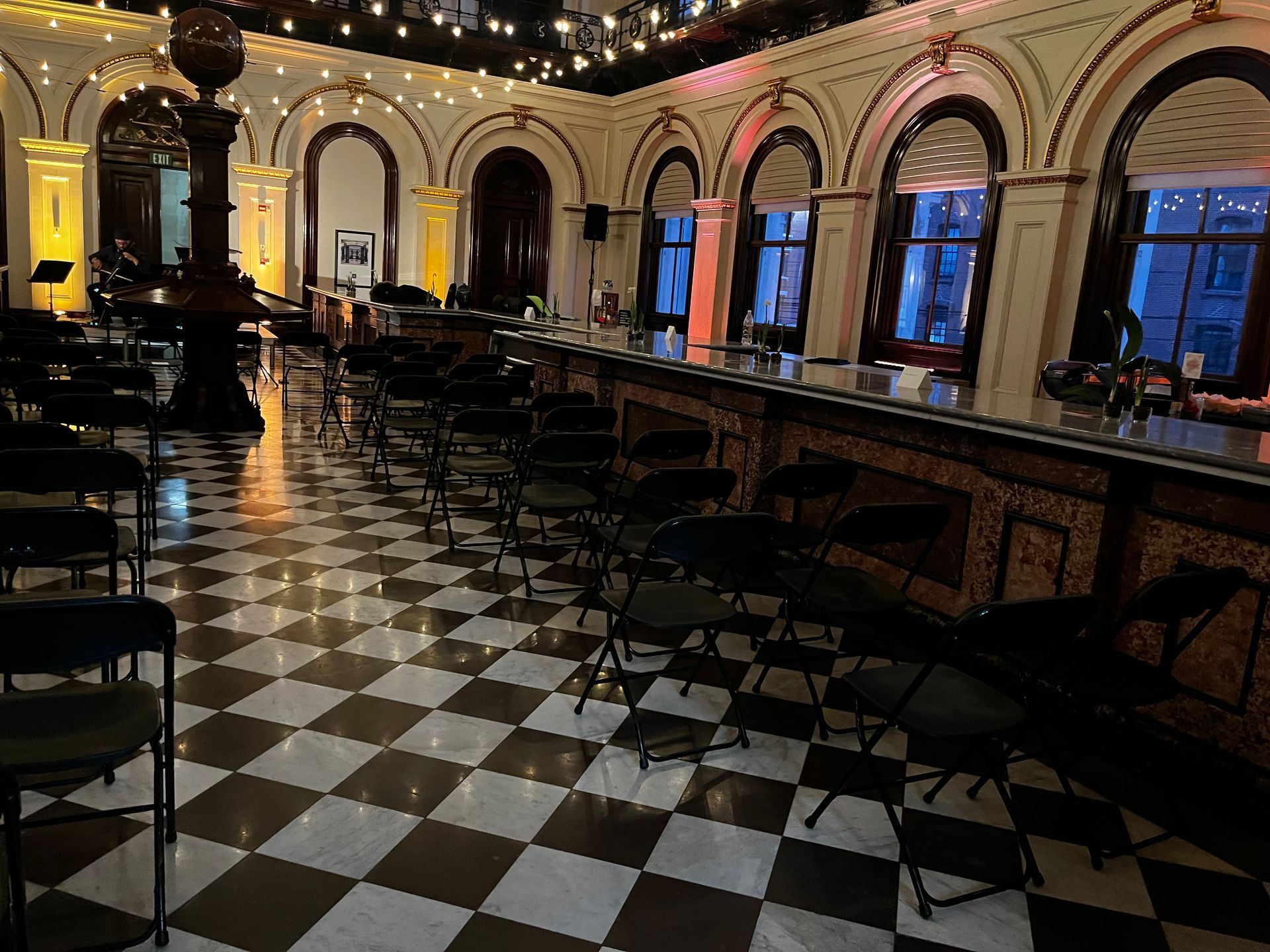 Elegant room with checkered floor, rows of chairs, bar, and arched windows.