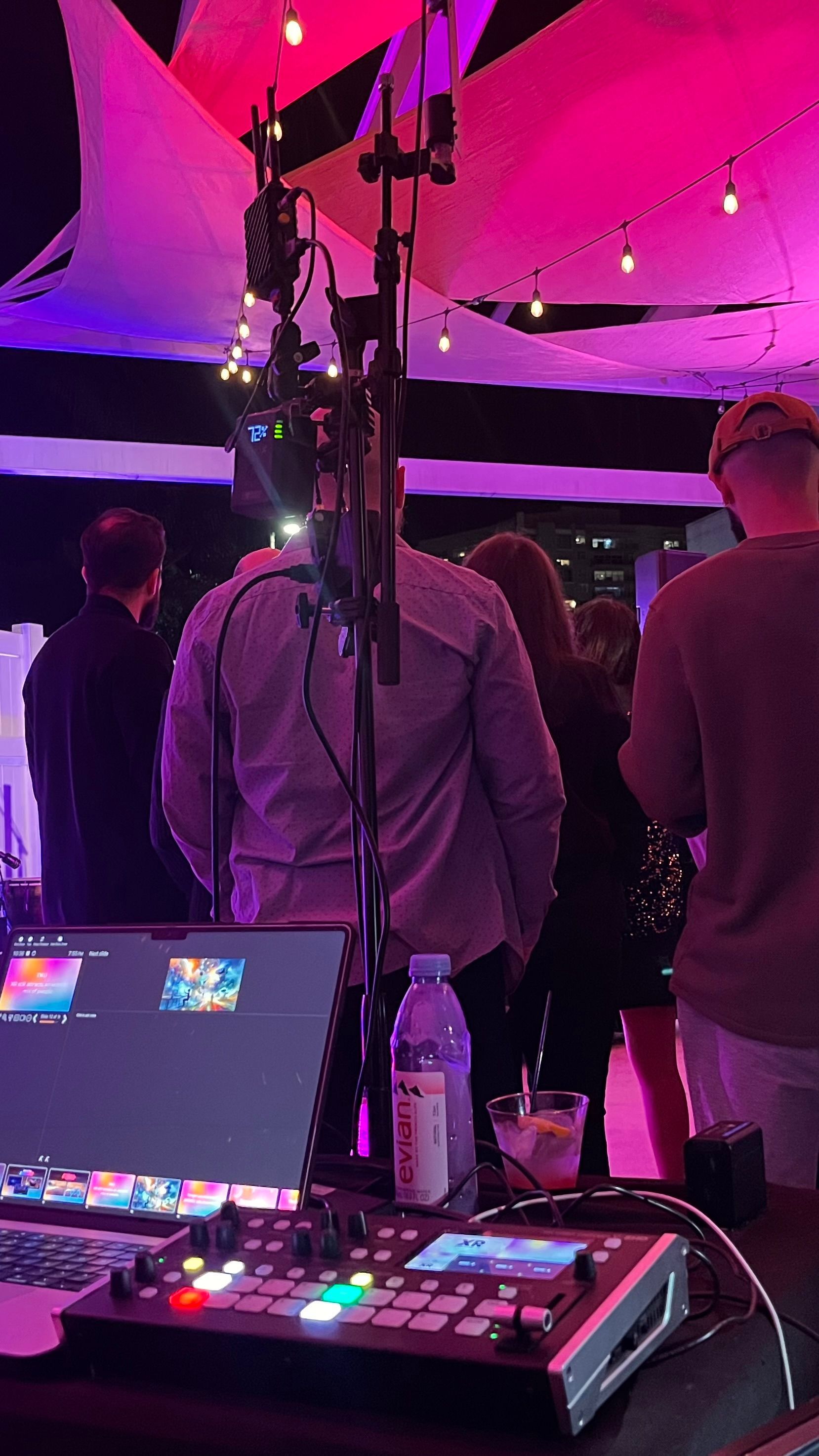 Video production setup at an outdoor event. People stand near a screen and equipment under pink and white lights.