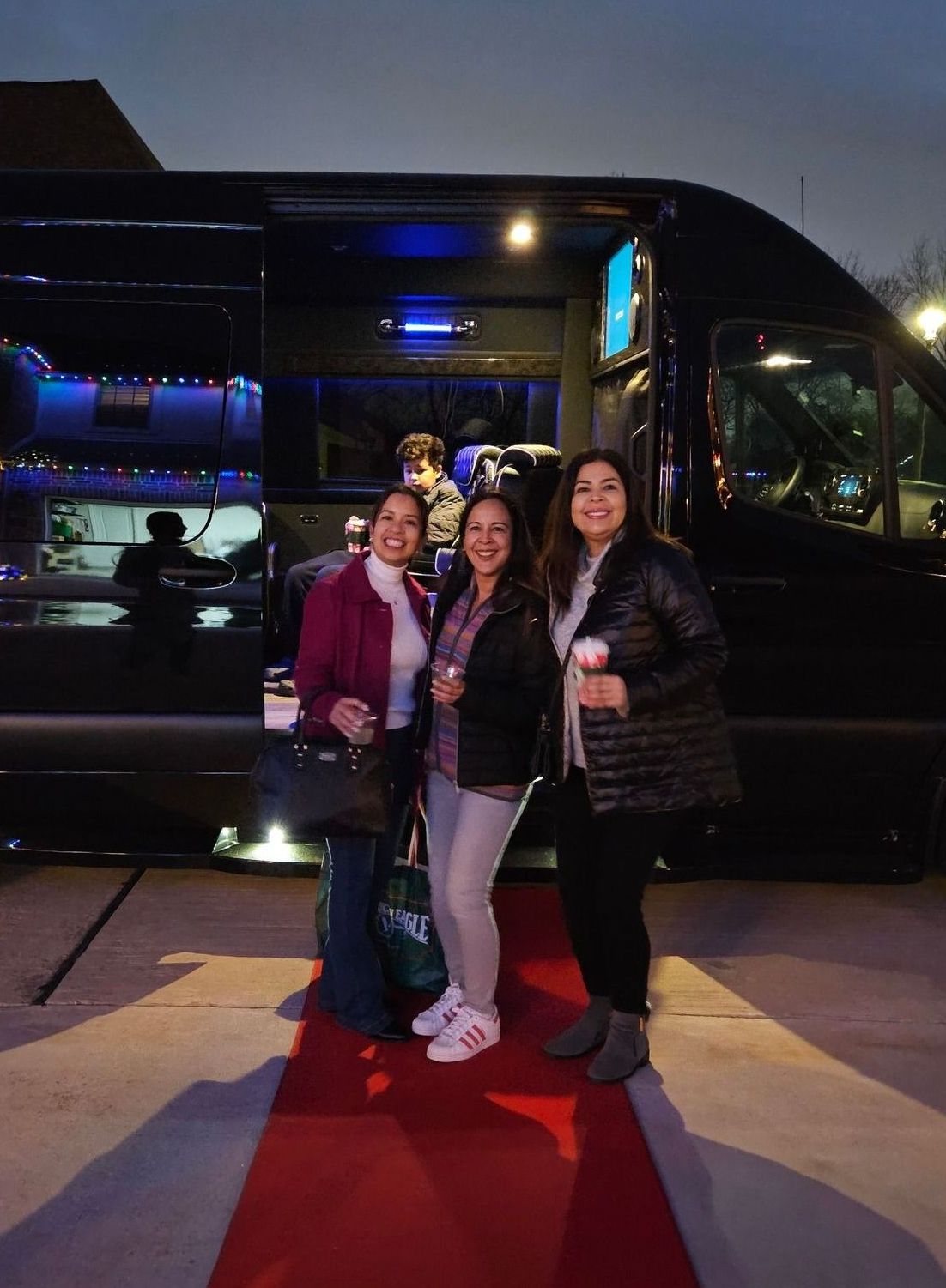 Three women are standing in front of a black van on a red carpet.