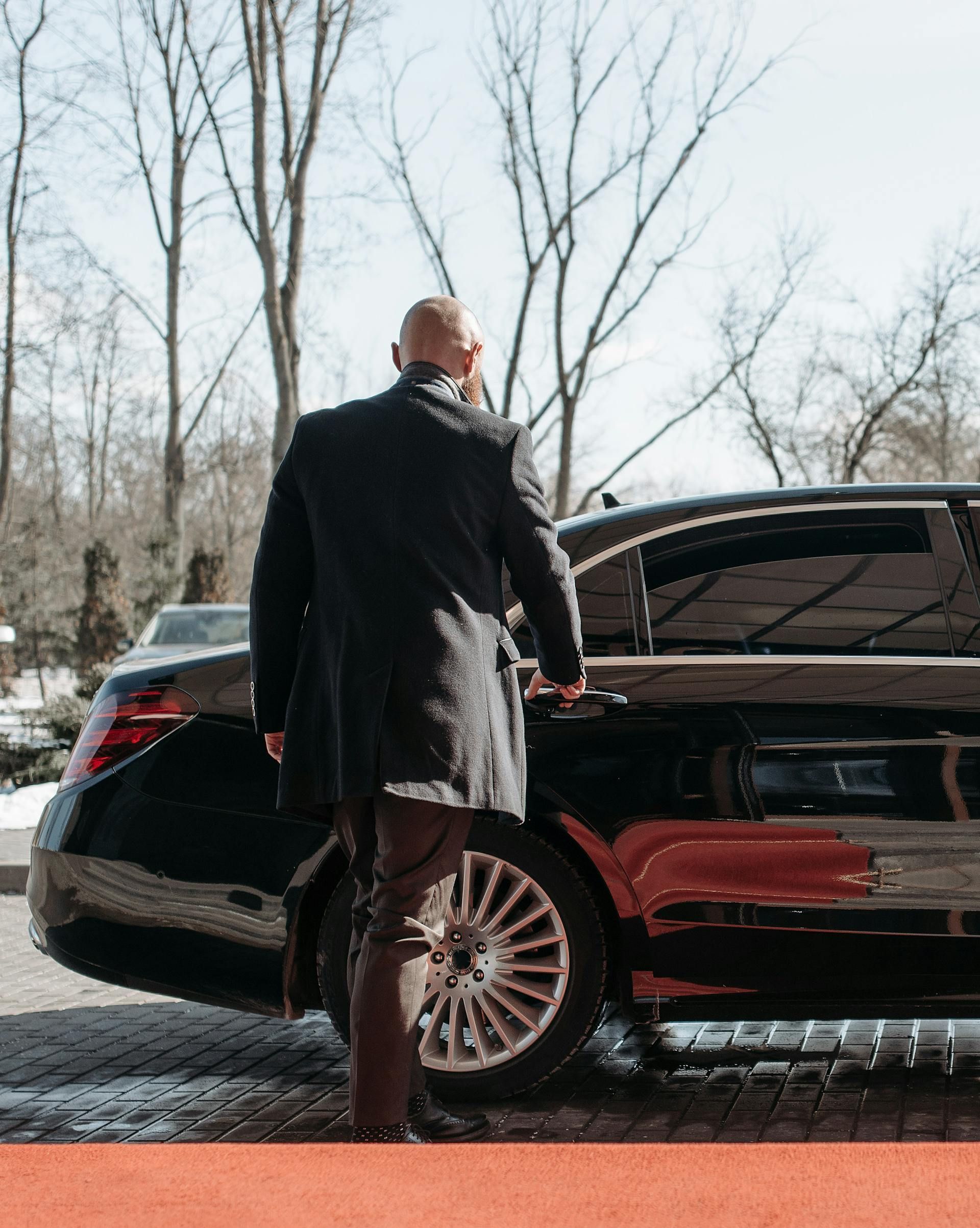A man in a black coat is opening the door of a black car.
