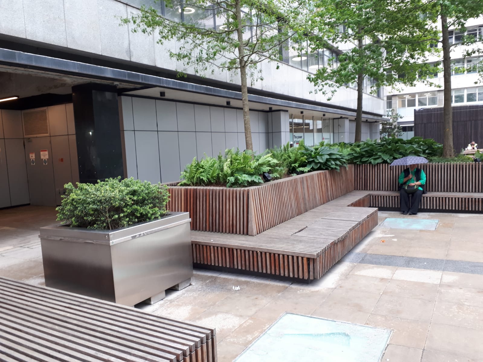 Outdoor seating area with wooden benches and planters, under a building with a person holding an umbrella.