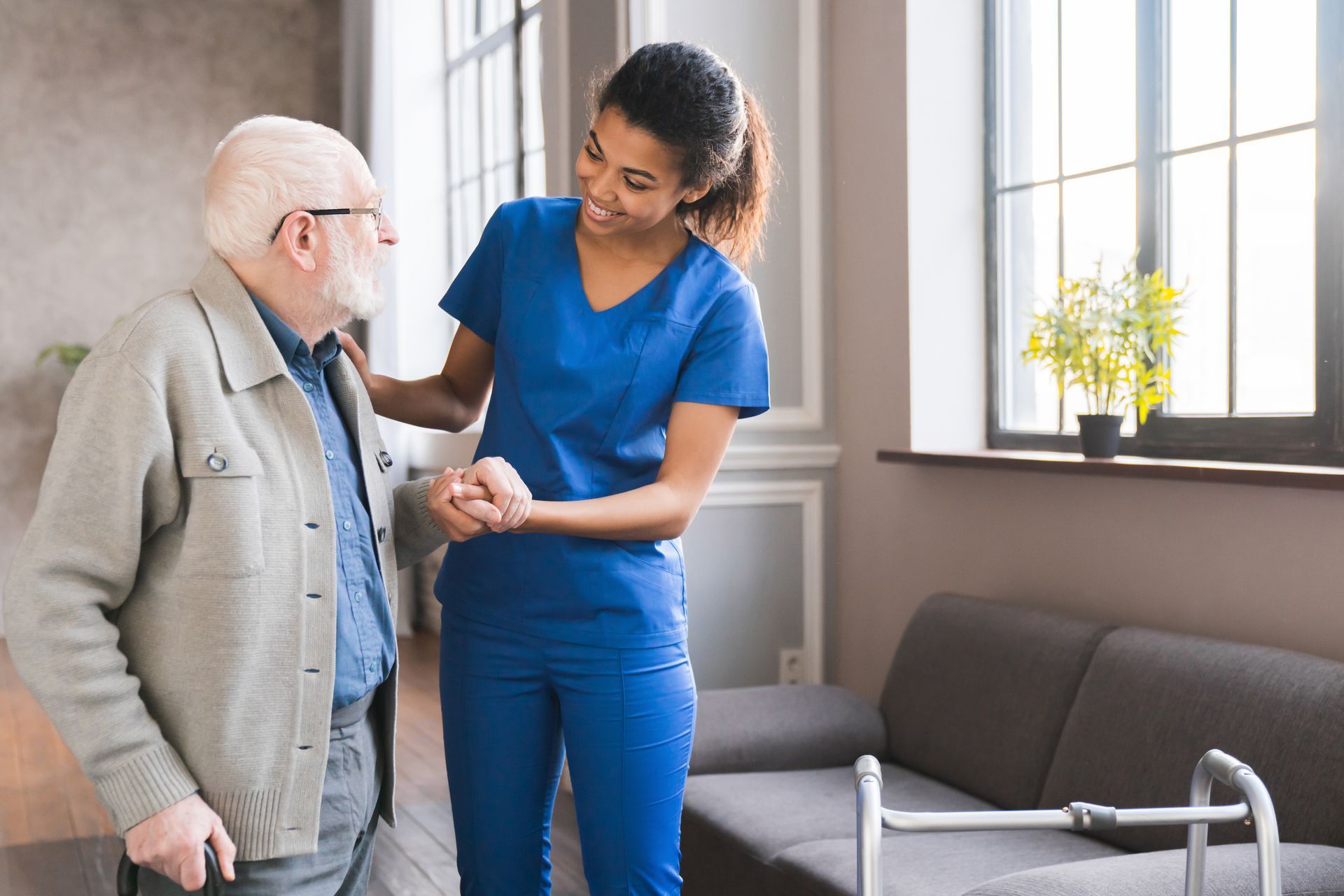 Caregiver helping an older person walk near a sofa and a walker.