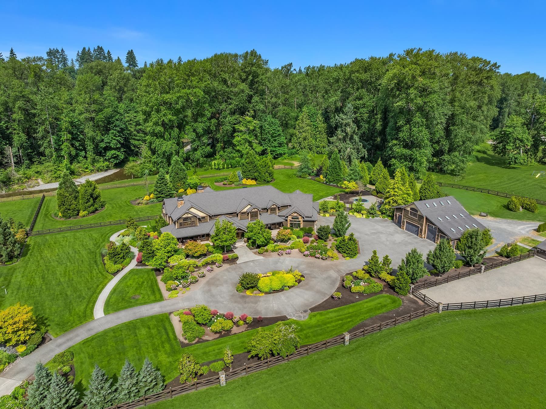 Aerial view of a large, luxurious home with a circular driveway, lush gardens, and a separate garage/stable area, surrounded by green fields and trees.