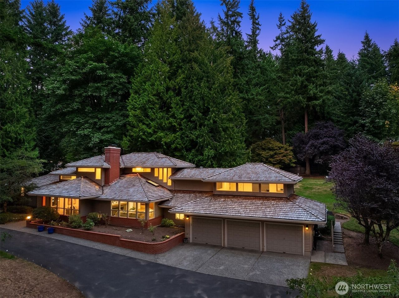 A large, multi-level home with a dark shingle roof, set in a lush, forested area at dusk.