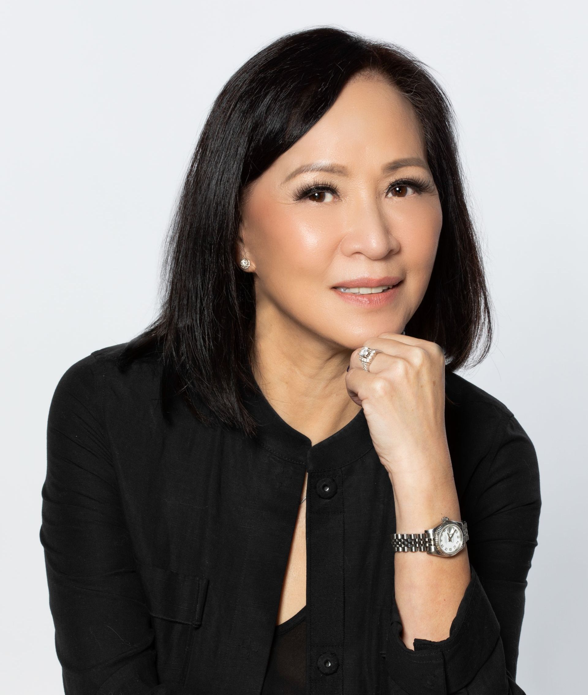 A woman wearing a black jacket and a watch is sitting with her hand on her chin.