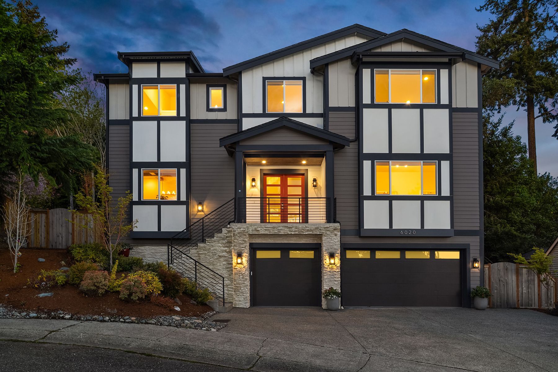 Modern three-story house at dusk with lit windows, stone accents, and two garage doors