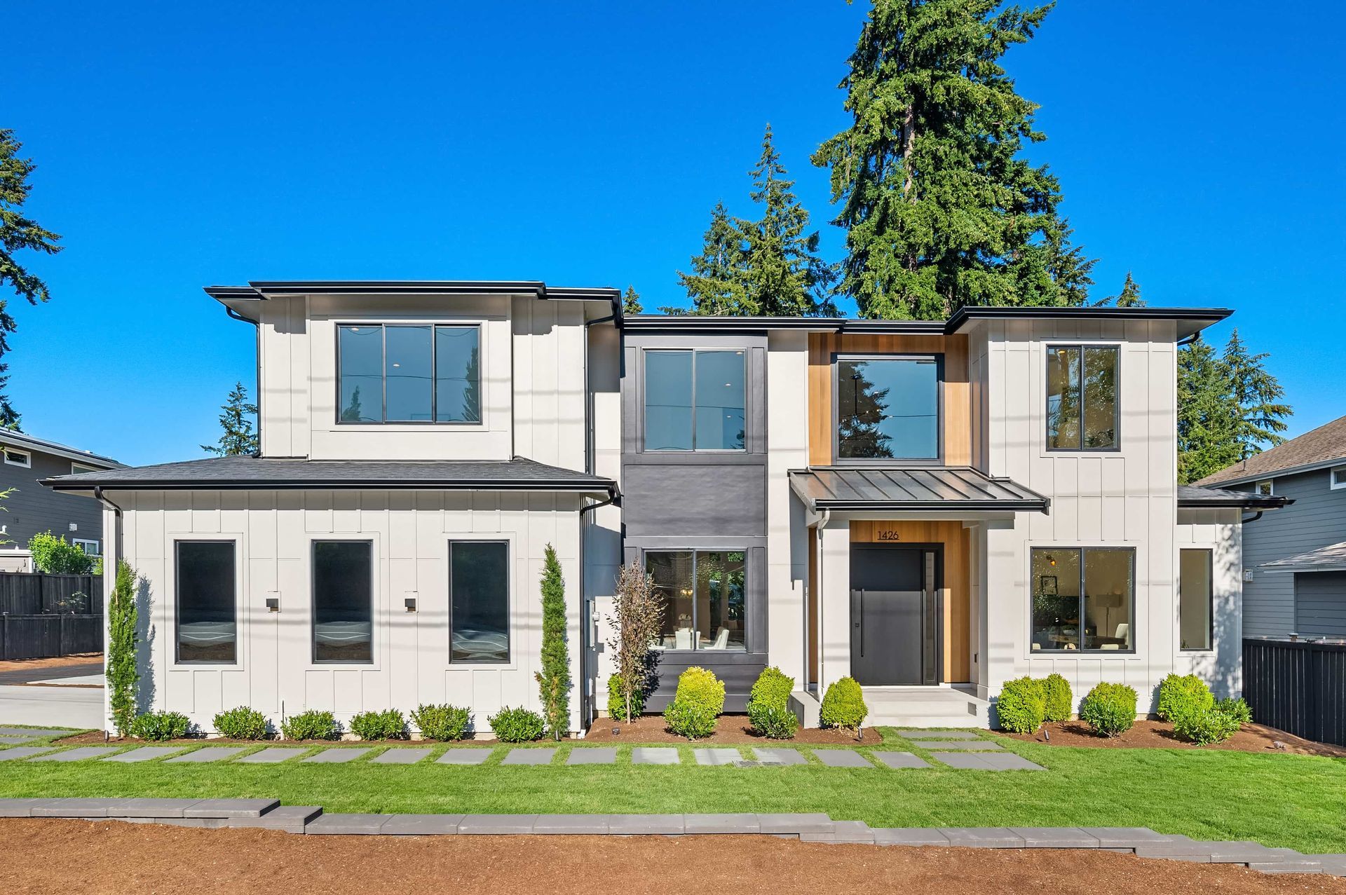 Modern two-story house with a light exterior, dark windows, and a black door. Green lawn and landscaping in front.