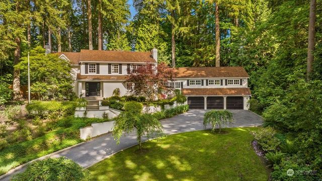 Two-story white house with black shutters and a three-car garage, set amongst tall green trees and lush landscaping.