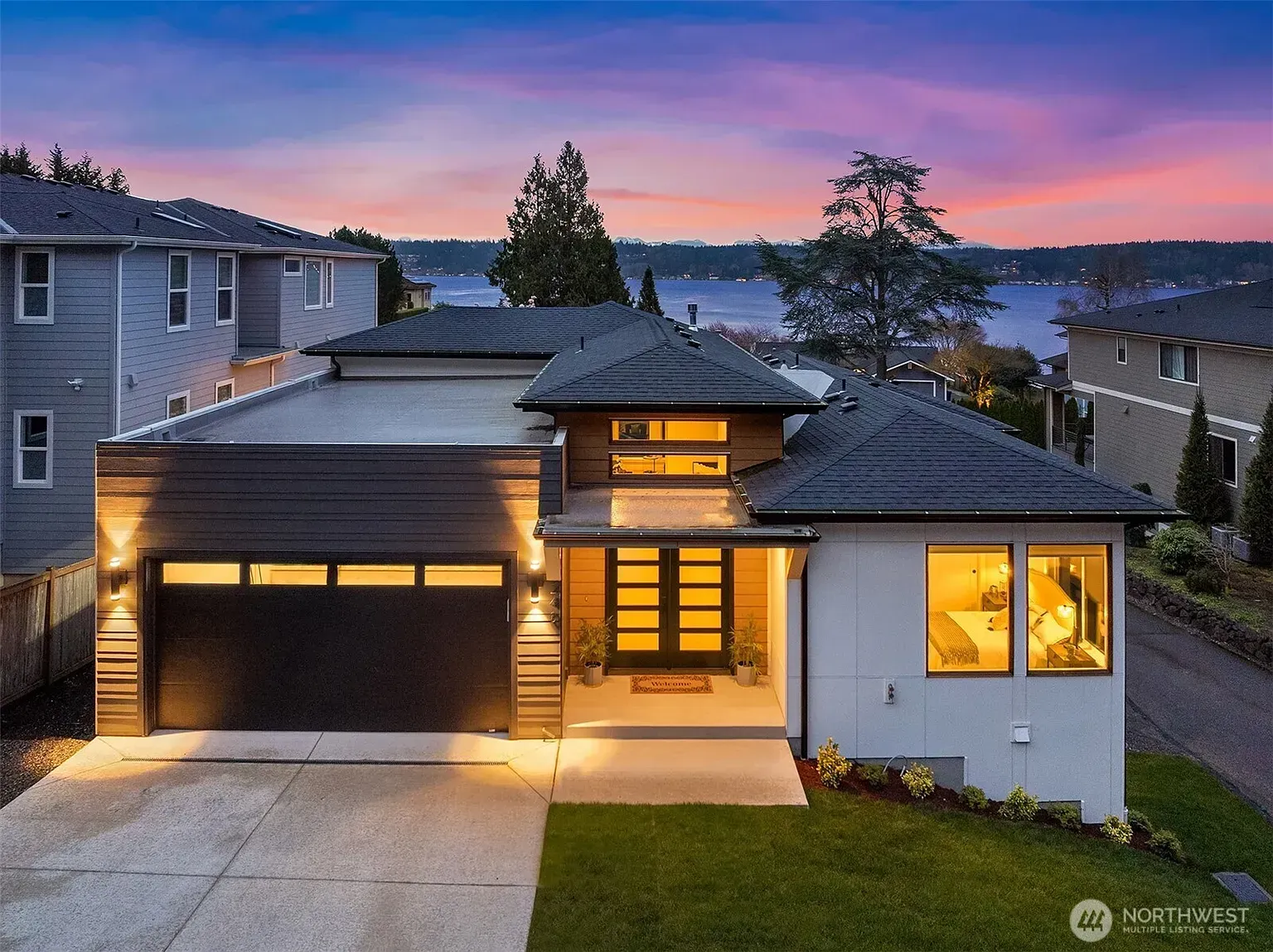 Modern multi-story house with stone and wood exterior, driveway, and tiered landscaping at dusk.