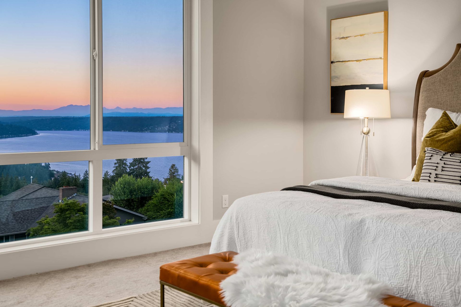 Bedroom with a large window overlooking a lake at sunset. Features a bed with neutral bedding, a bench, and artwork.