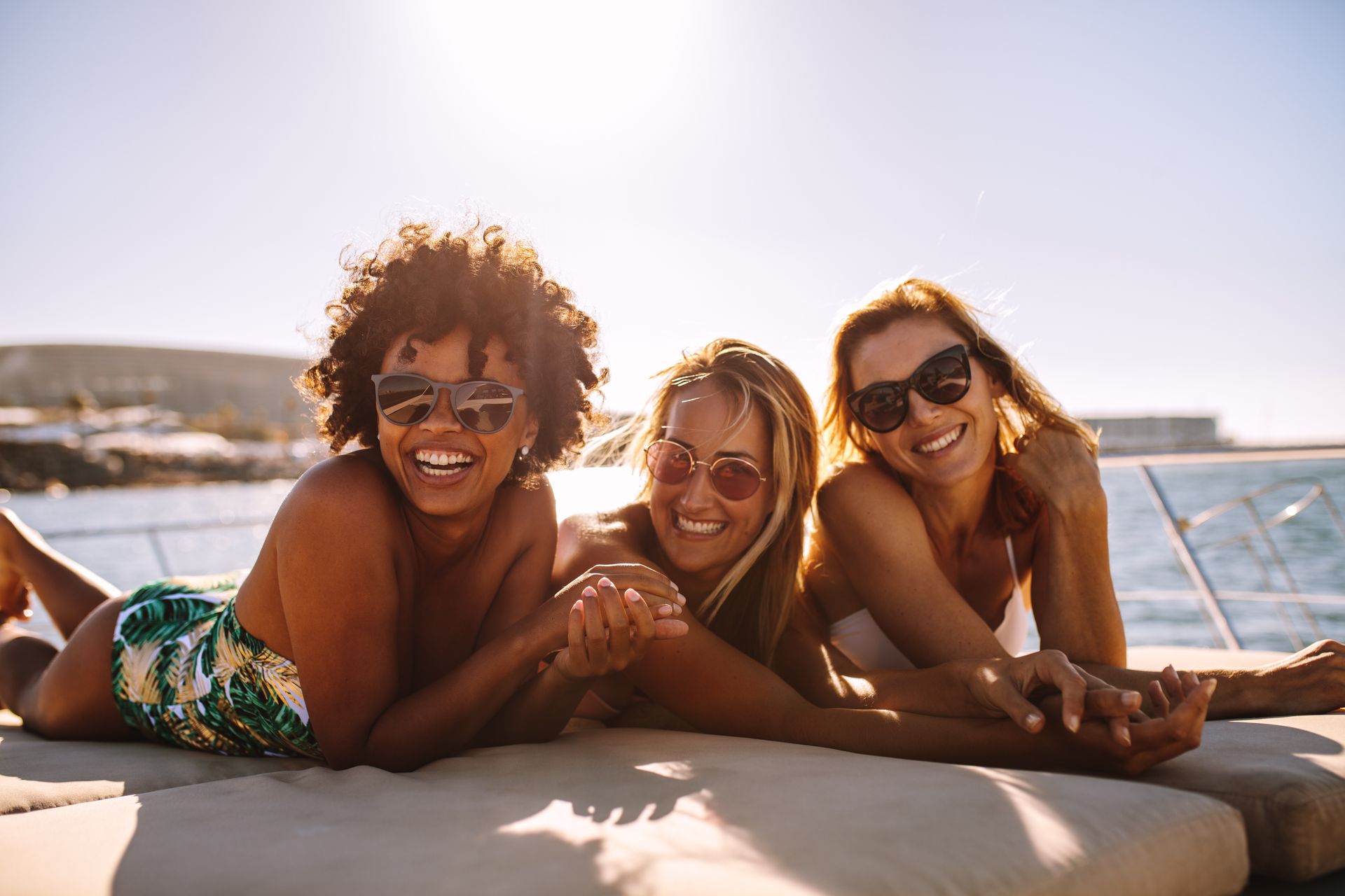 Three friends smiling on a boat deck, sunbathing and holding hands.