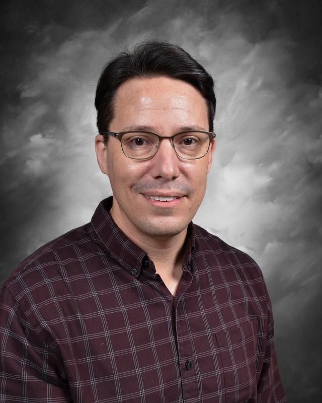 A man wearing glasses and a jacket is sitting in front of a blue background.