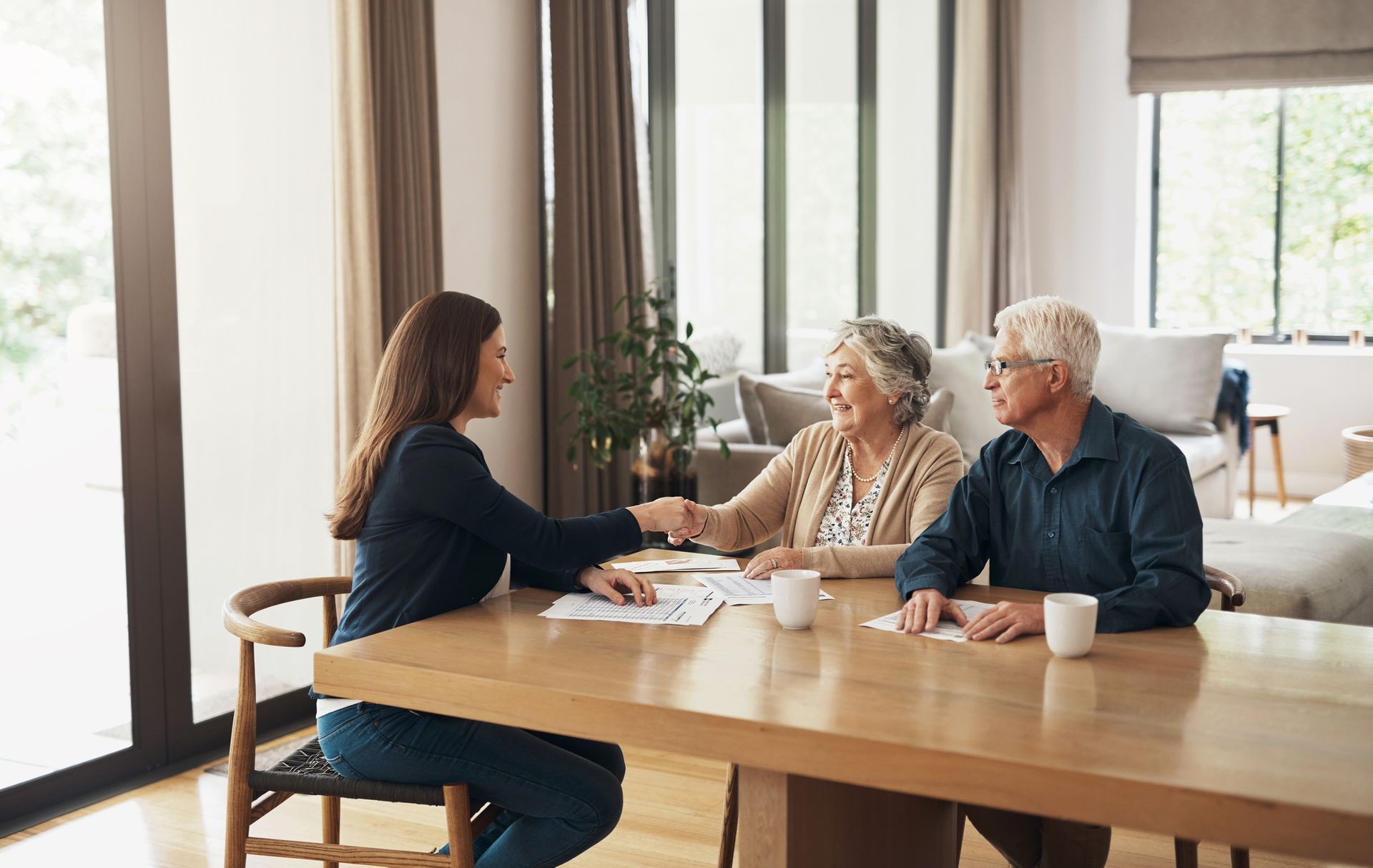 Woman shaking hands with an elderly couple at a table, documents and coffee cups present in a home s