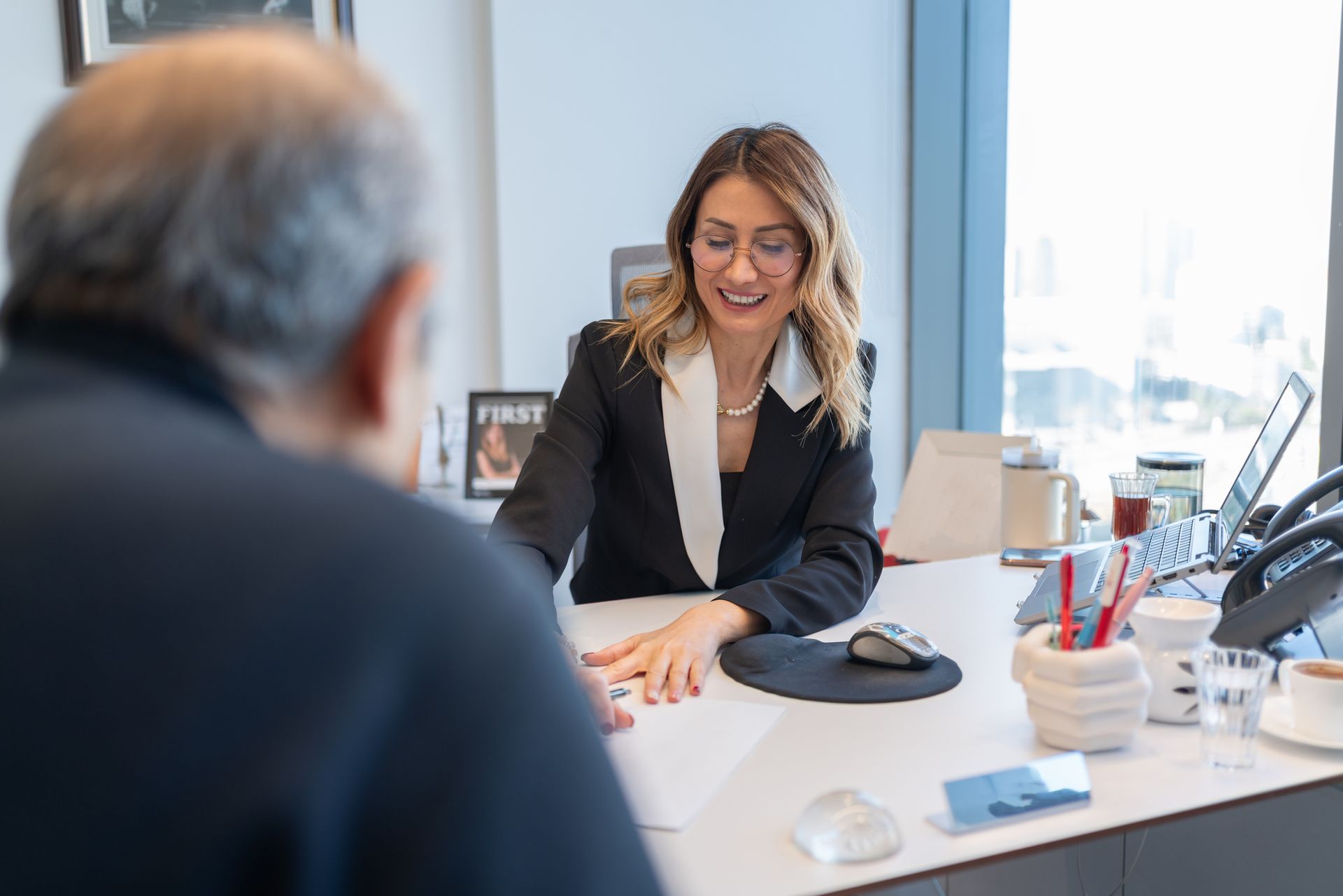 Woman in blazer smiles, leans over desk, talking to a man out of focus. Office setting, paperwork.
