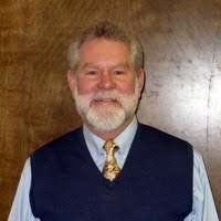 Man with a white beard and a navy vest, smiling, against a brown background.