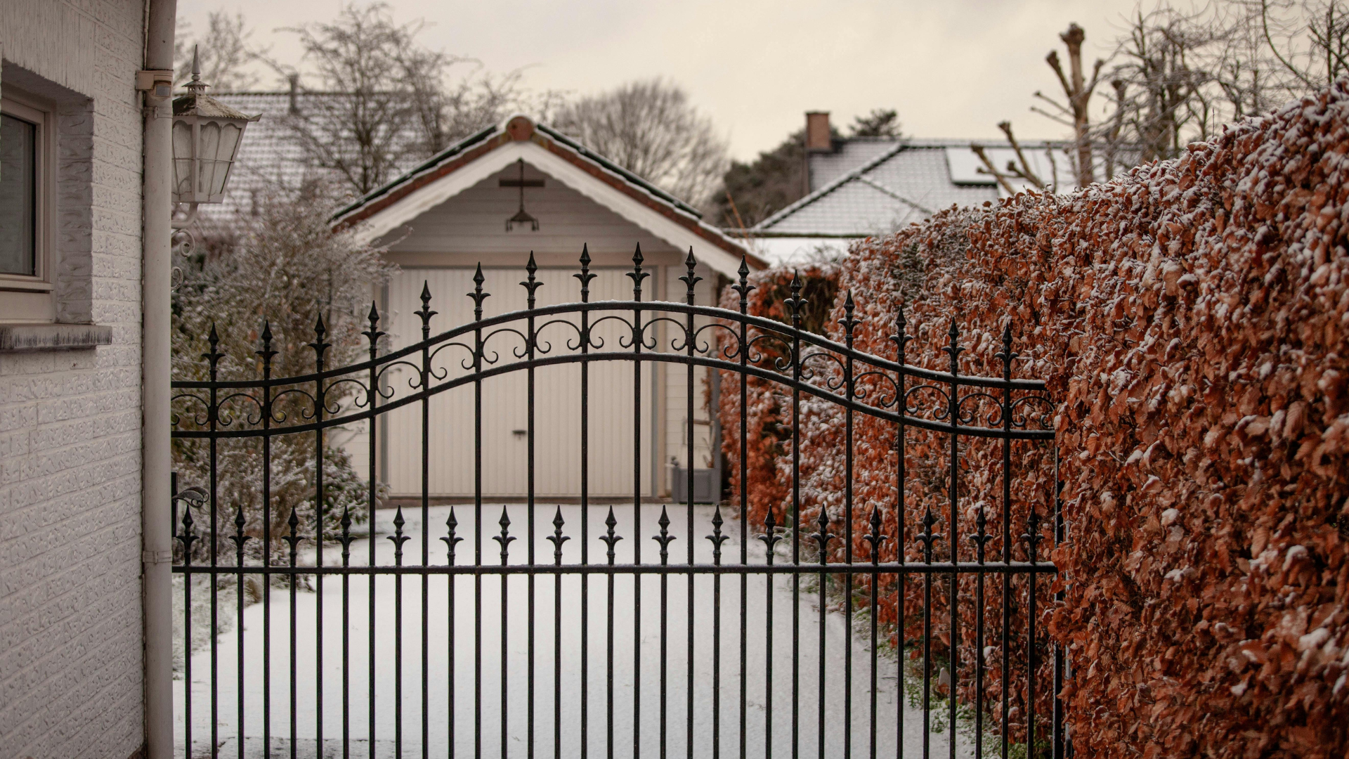 A black metal gate stands before a garage driveway covered in snow, bordered by a hedge with brown, winter-worn leaves.