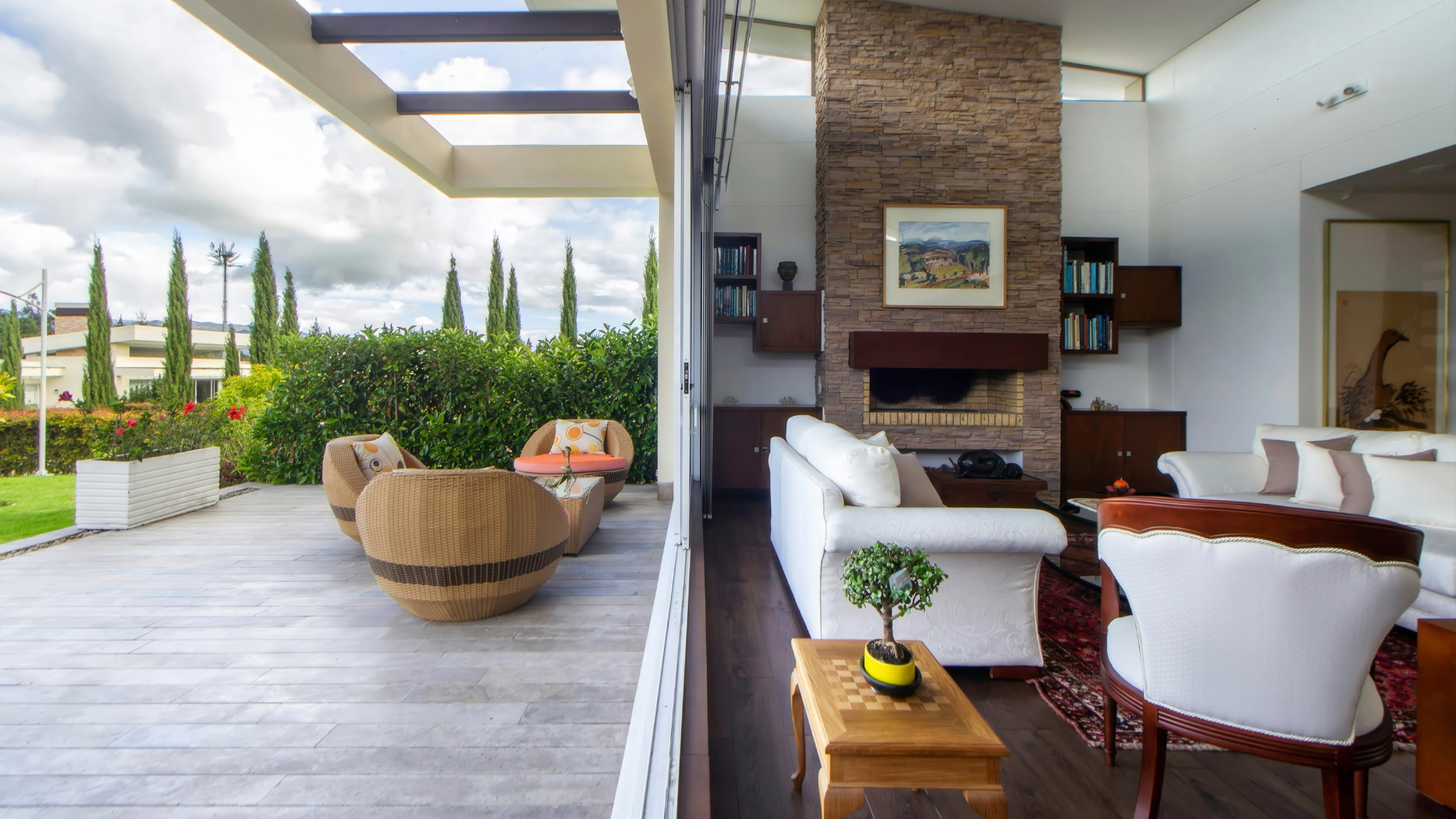 A bright, modern indoor-outdoor living space with white sofas, a stone fireplace, and a patio with wicker lounge chairs.