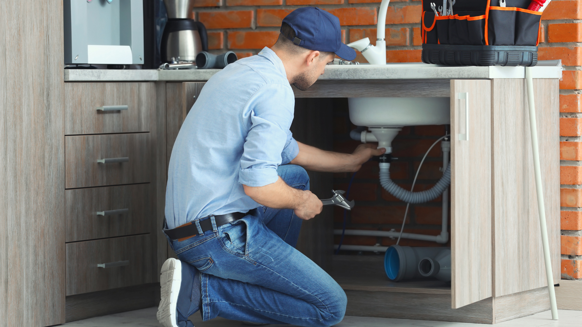 A plumber in a blue shirt and cap kneels in a kitchen to repair the drain pipes under a sink.