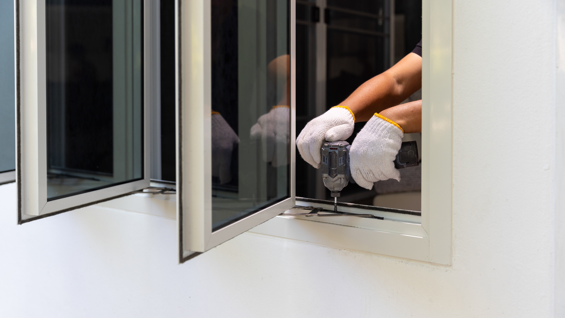 A person wearing white gloves uses a cordless drill to install or repair a white framed window.
