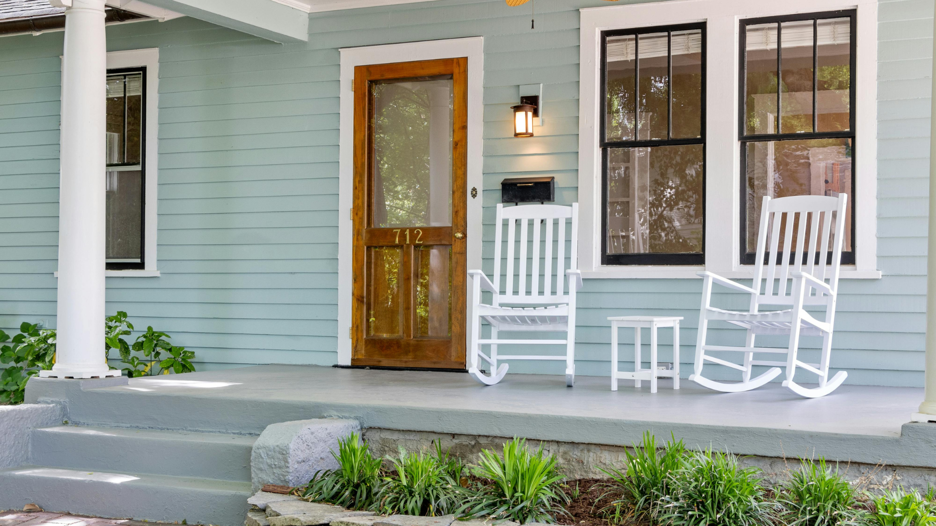 Light blue porch with two white rocking chairs and a small table, featuring a wooden door and two black-framed windows.