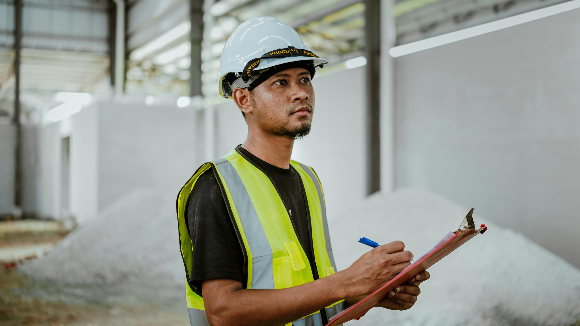 A person in a hard hat and high-visibility vest stands in an unfinished building, holding a clipboard and pen.