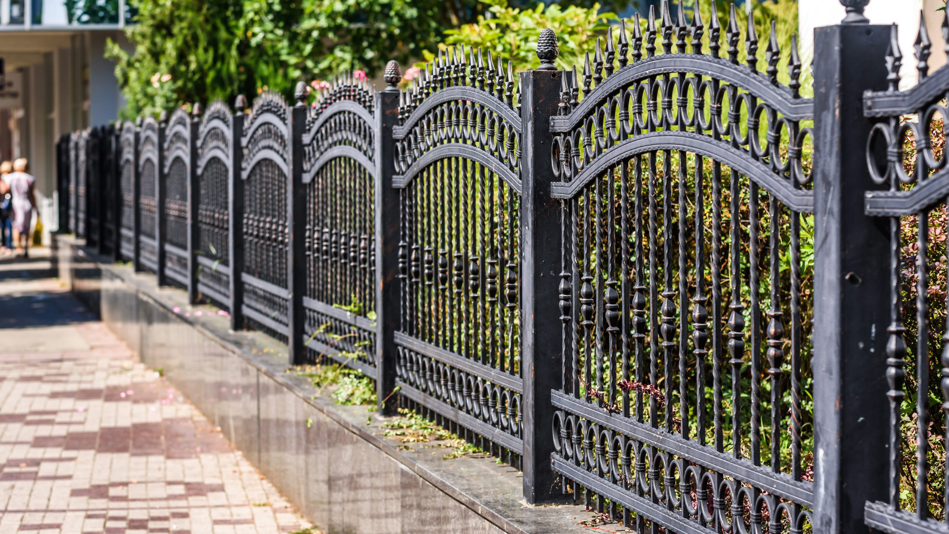 A black wrought-iron fence with arched decorative tops lines a paved sidewalk next to greenery.