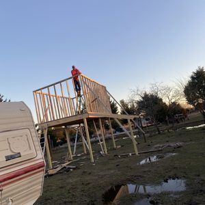 A man is standing on top of a wooden structure in a field next to a trailer.