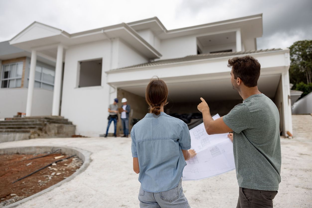 Couple holding blueprints while looking at a white modern house under construction.