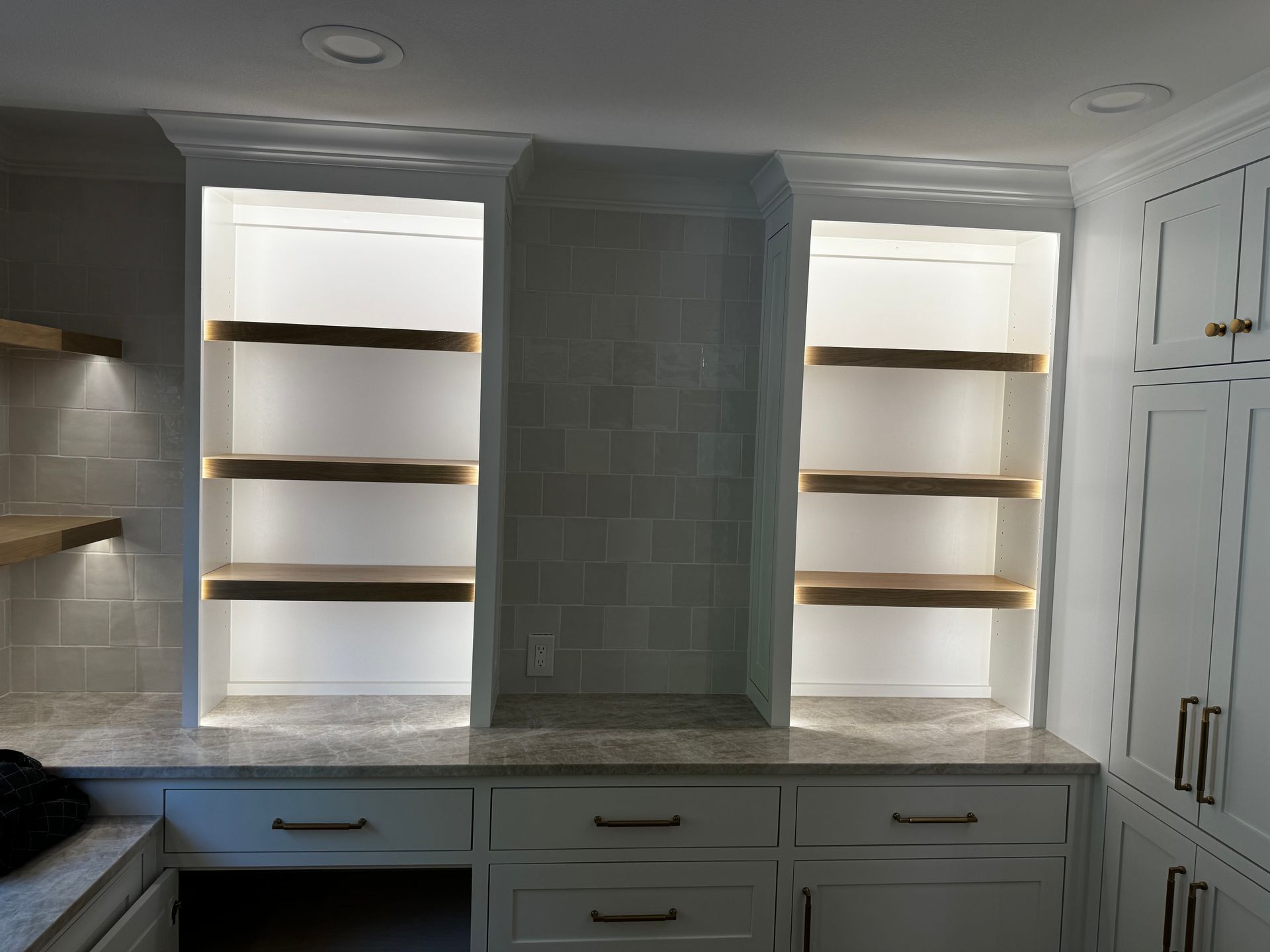 Built-in kitchen cabinetry with white shelves, gray backsplash, and marble countertop under soft lighting