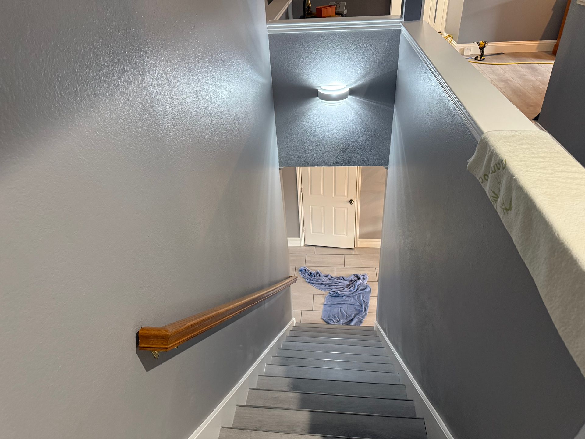 Stairwell with gray walls, wooden handrail, and steps leading down to a lit basement area.