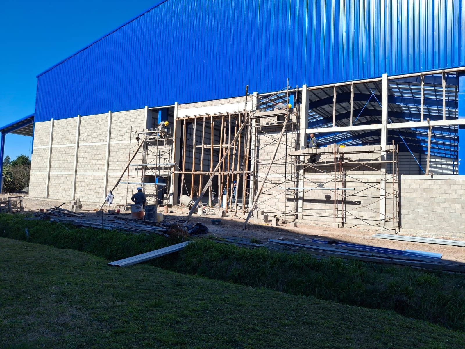 Obra en construcción con techo de metal azul, paredes de bloques grises, andamios y obreros.