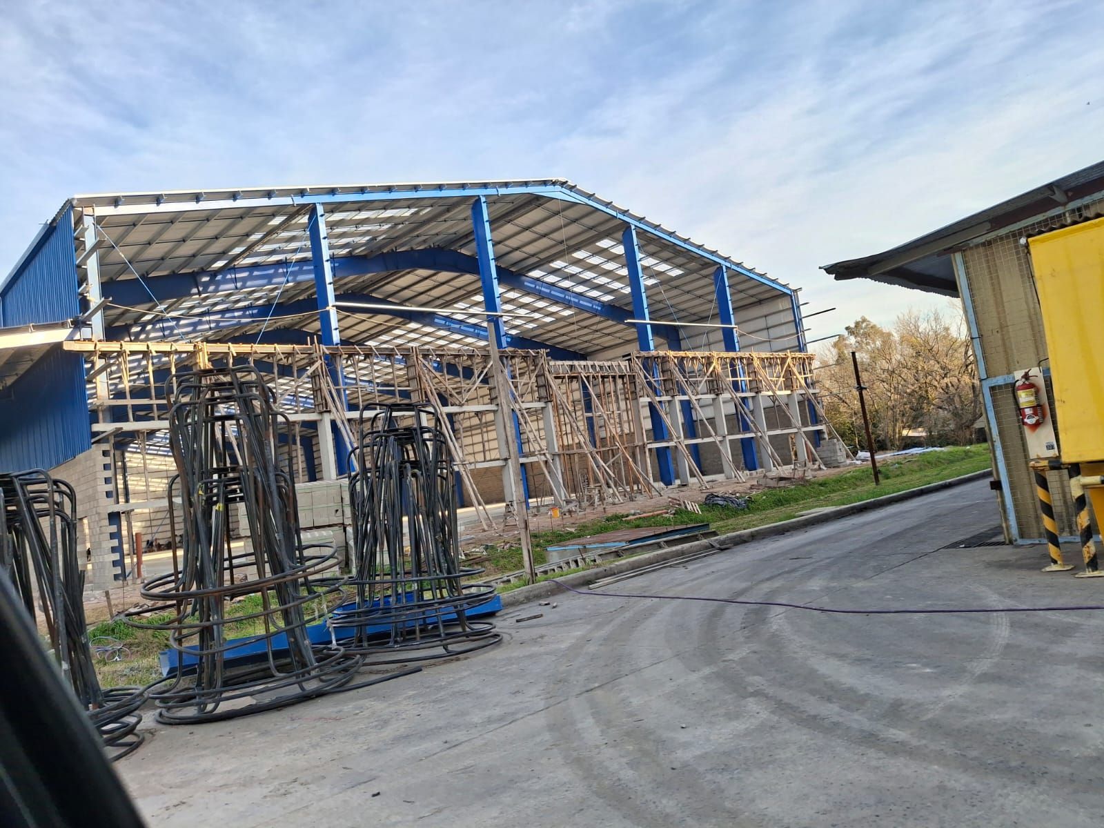 Exterior de almacén con techo azul, construcción visible, varillas de acero en primer plano.